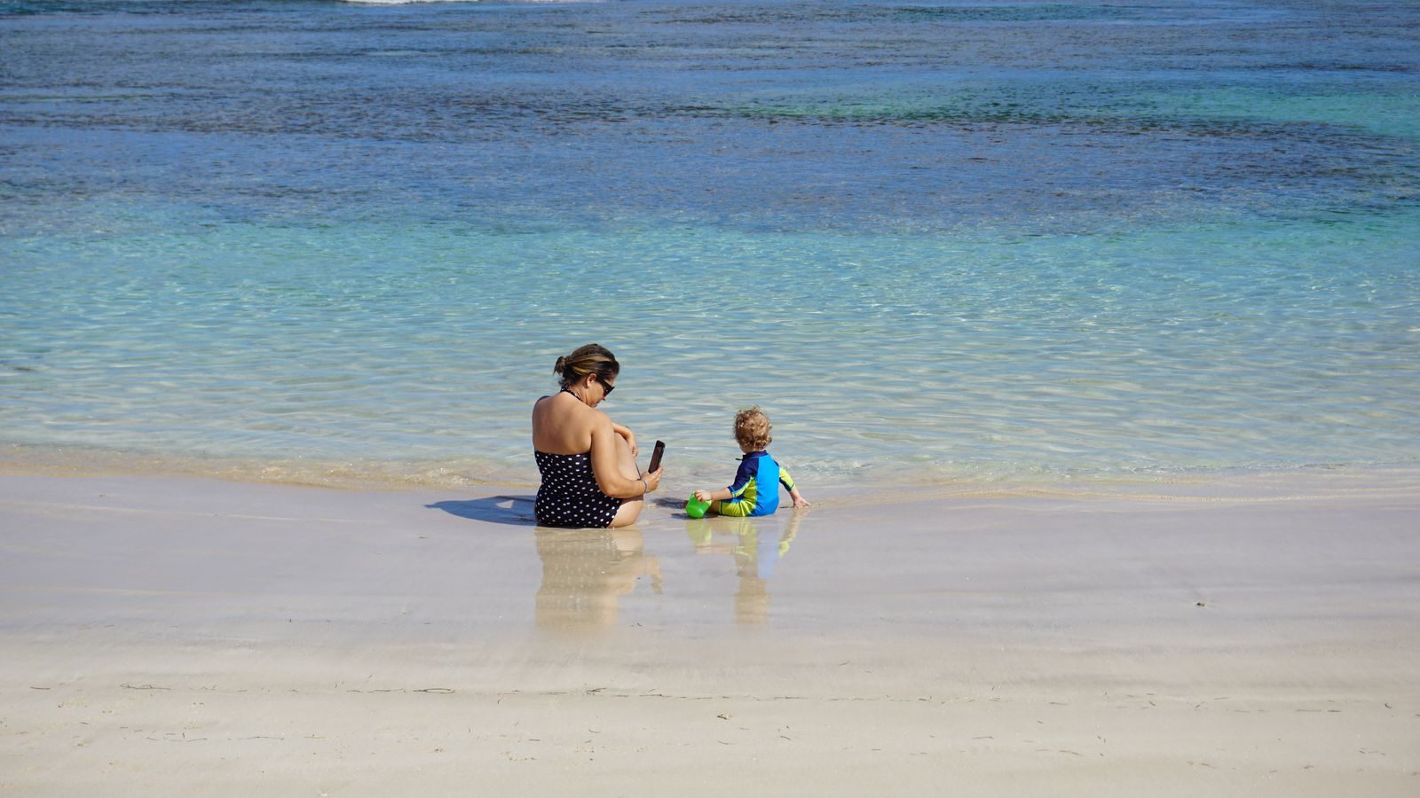 Mother and young child sitting together in shallow turquoise water, cruising with kids tips in action on a family cruise beach day